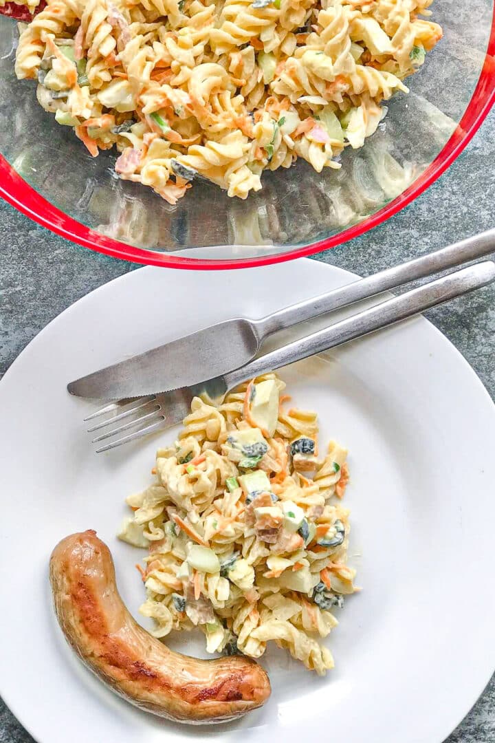 Pasta salad on a white round plate with a grilled sausage on the side