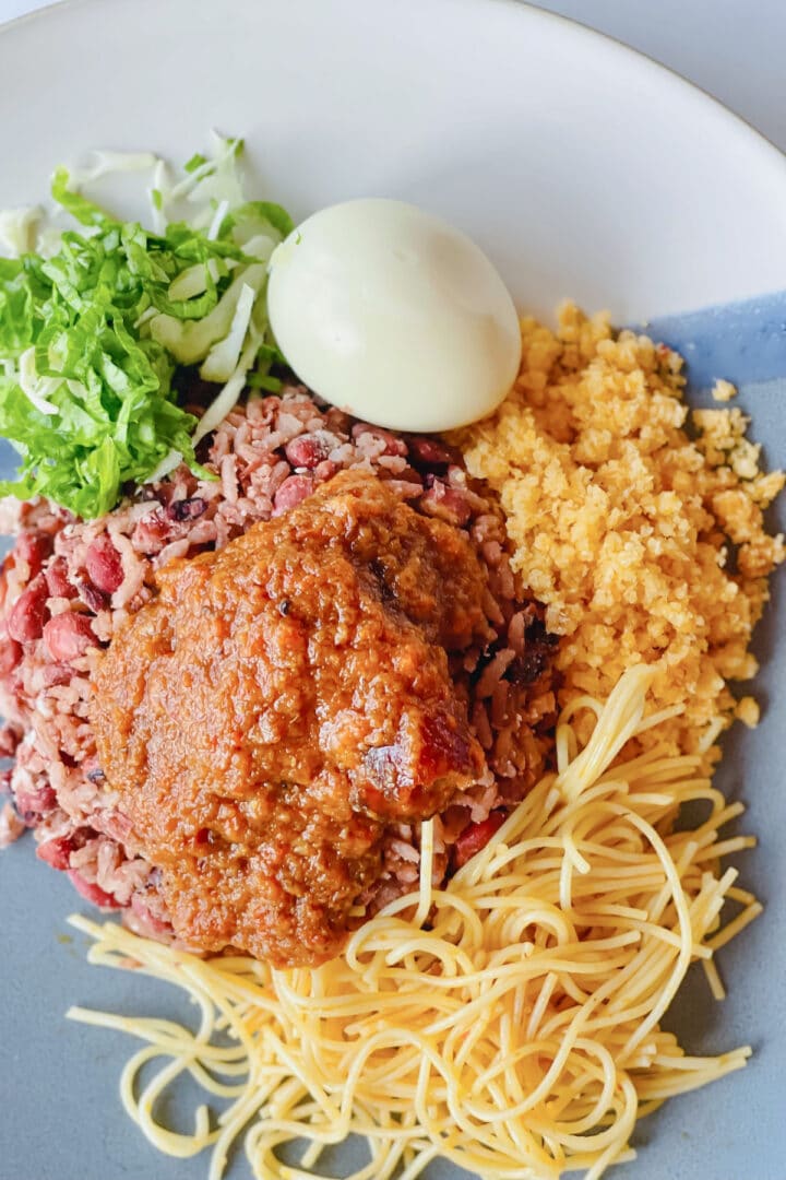 A plate of waakye and stew with a boiled egg, cut lettuce, gari and spaghetti.