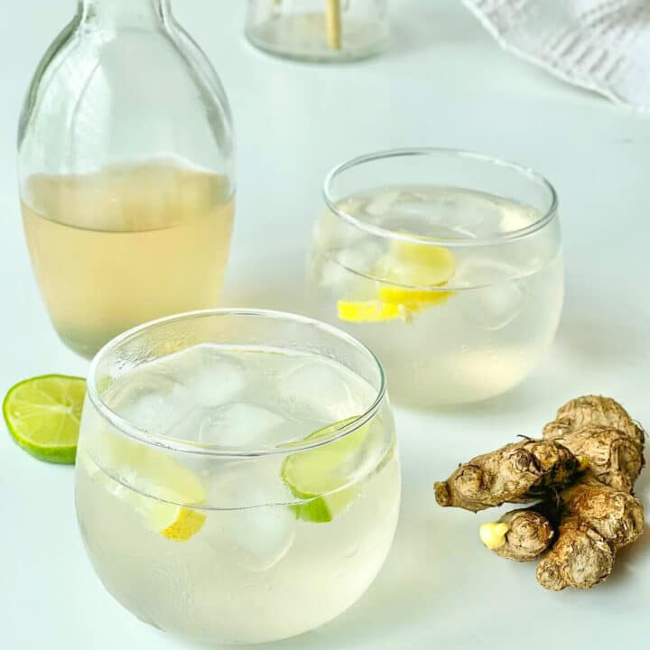 Two round short glasses of ginger ale and lime slices with ginger syrup in the background