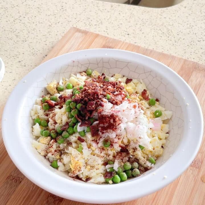 Chopped bacon, onions eggs, peas in a white round plastic bowl.