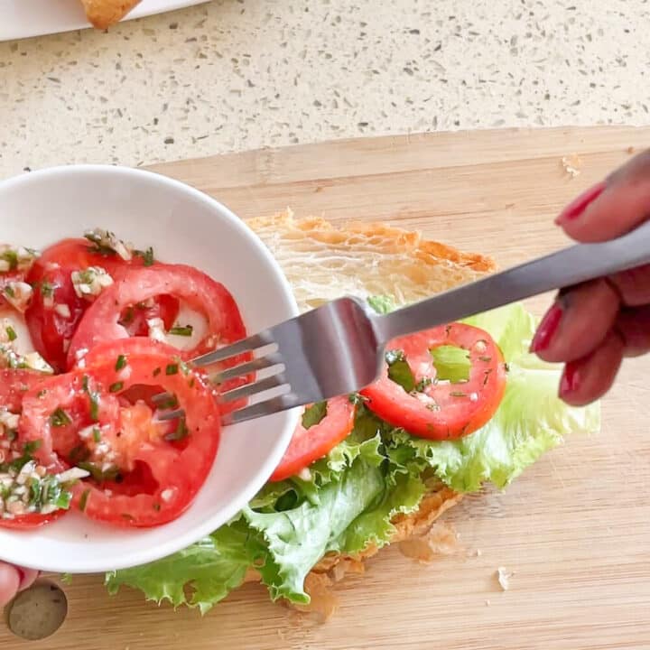 Marinated sliced tomatoes in a white bowl with a couple on a croissant on a wooden board.