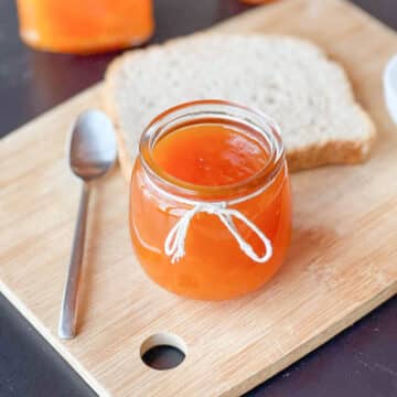 Mango hibiscus jam in a small glass jar with a slice of bread in the background.
