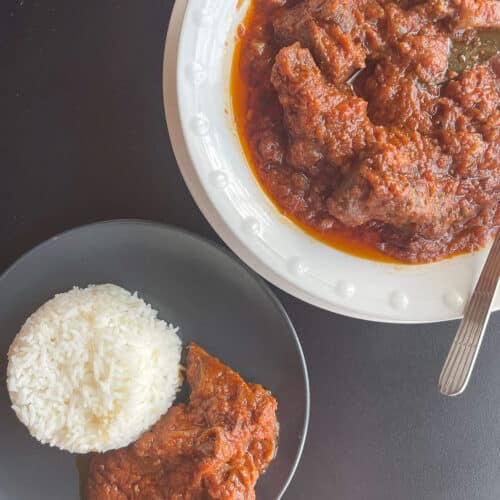 Goat meat stew on a black plate with a heap of boiled plain rice. A white bowl of goat meat stew in the top right corner.
