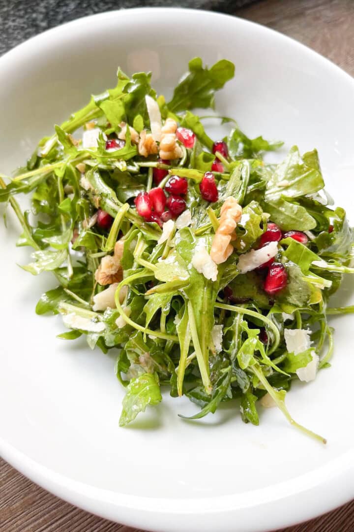 Arugula salad with pomegranate seeds, parmesan shavings, and chopped walnuts in a white bowl.