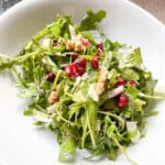 Arugula salad with pomegranate seeds, parmesan shavings, and chopped walnuts in a white bowl.
